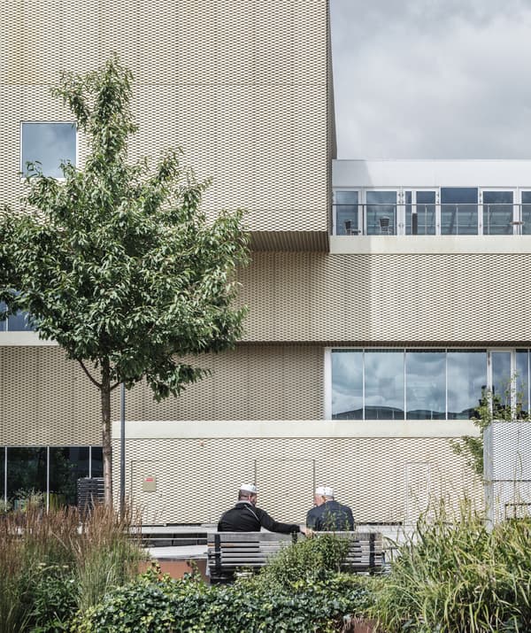 Deux hommes assis sur un banc dans un jardin à l'extérieur d'un bâtiment habillé de métal doré.