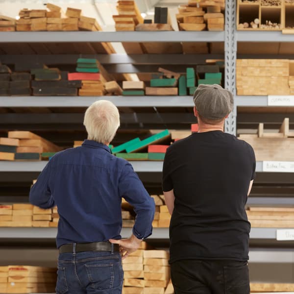 Deux hommes, l’un habillé de bleu et l’autre de noir, se tiennent côte à côte devant un assortiment de bois d’œuvre dans un entrepôt.
