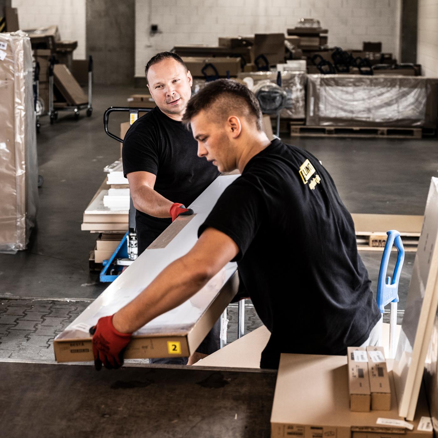 Deux hommes vêtus de t-shirts noirs et de gants de travail rouges chargent des paquets plats IKEA dans un camion.
