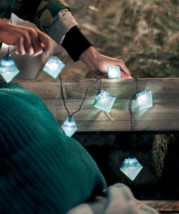 Deux mains décorant un banc public avec une guirlande lumineuse SOLVINDEN. Coussins et plaid sur le banc.