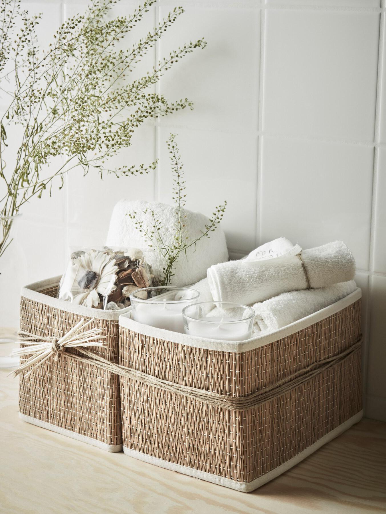 Deux paniers en paille, sur un fond en carrelage, liés l'un à l'autre par plusieurs ficelles nouées, et customisés avec des serviettes de bain, deux bougies en verre et un pot pourri.