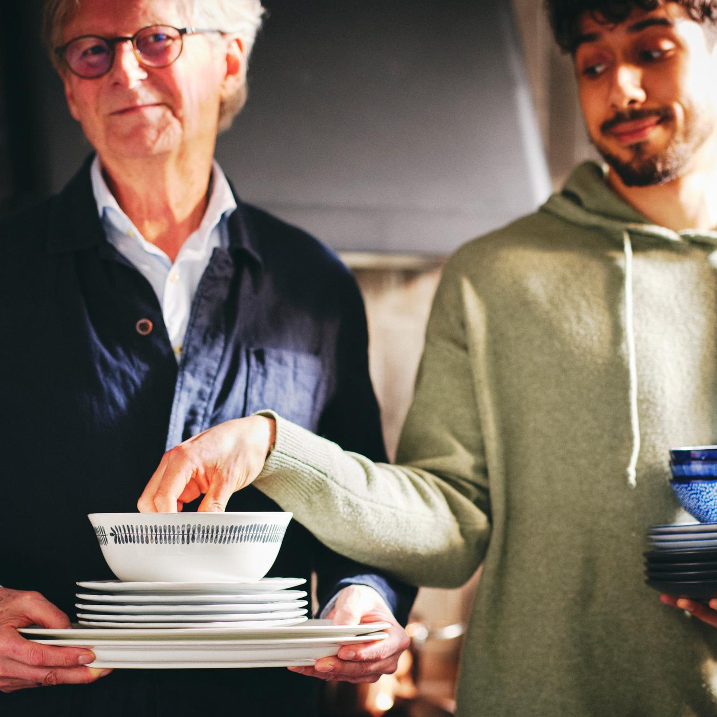 Deux personnes dont l’une tient une pile de plats et d’assiettes UPPLAGA blancs et un bol FRIKOSTIG blanc à motifs.
