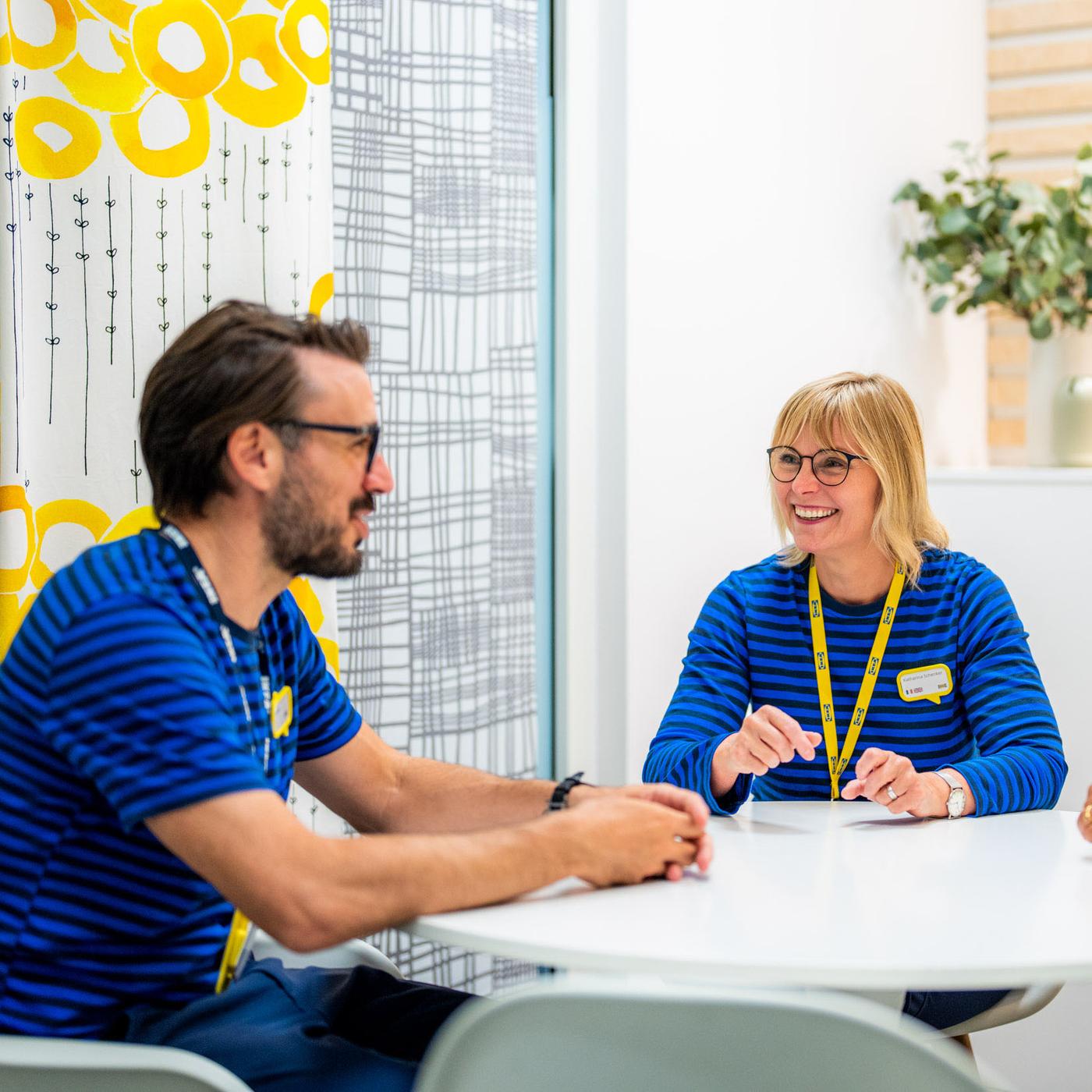 Deux personnes en uniformes rayés bleus discutent autour d'une table ronde moderne, dans un intérieur lumineux décoré de plantes.