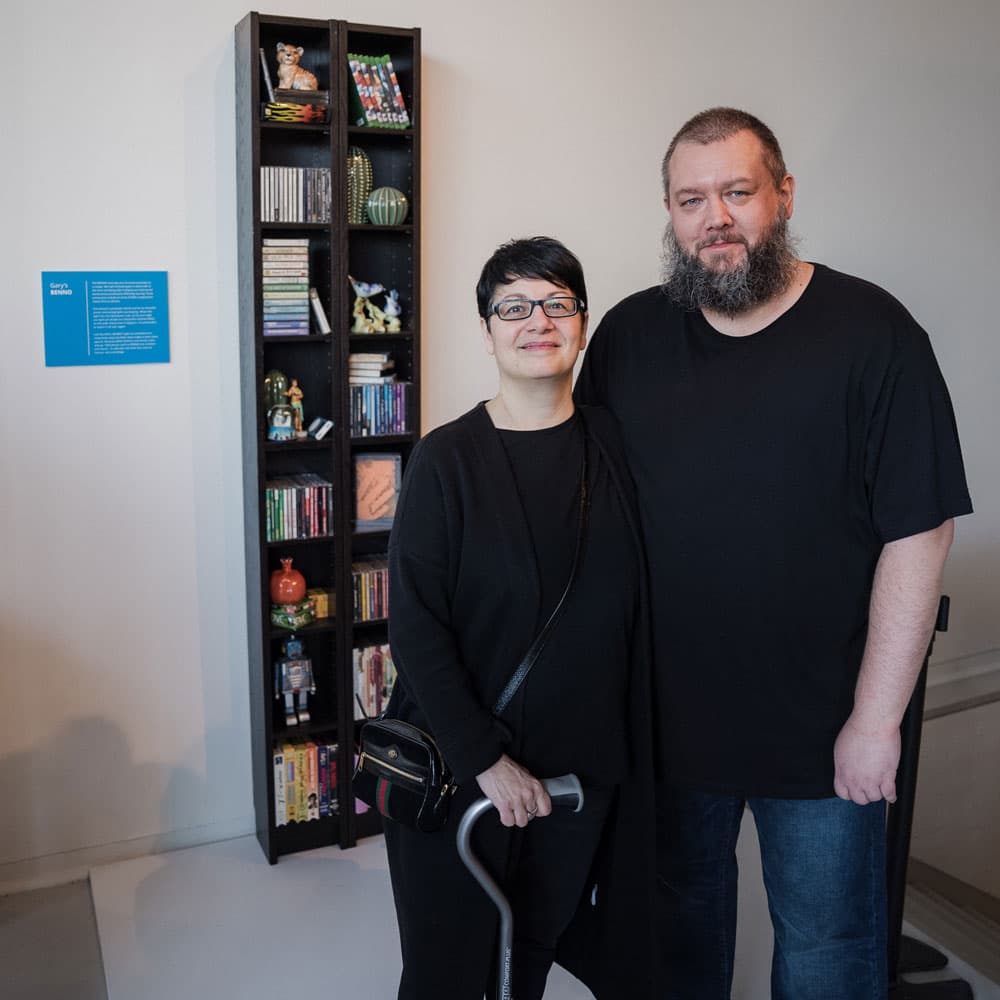 Deux personnes posent devant une bibliothèque IKEA BENNO, contenant différents articles tels que des décorations, des livres, des jouets et des CD de jeux vidéo.