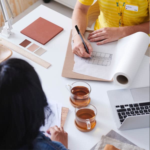 Deux personnes sont assises à une table, l'une dessine une carte sur du papier et l'autre observe, tandis que sur la table se trouvent deux tasses de thé, un ordinateur portable, des échantillons de couleurs, une règle et des documents, entre autres.