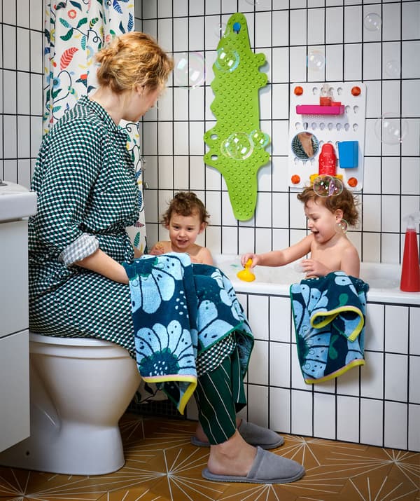 Deux petits enfants dans une baignoire à carreaux blancs. Une femme est assise sur le couvercle de la toilette juste à côté.