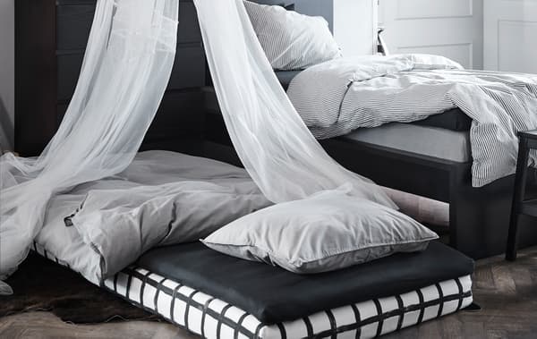 Deux petits matelas empilés et arrangés en lit sur le sol, à côté du lit. Un ciel de lit surplombe le lit d'appoint.