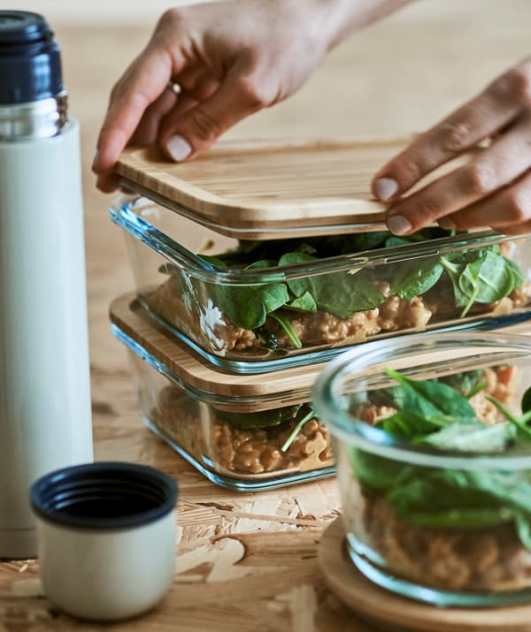 Deux piles de récipients alimentaires en verre et un pichet isotherme sur une surface en bois. Une main place un couvercle en bambou hermétique sur un récipient en verre.