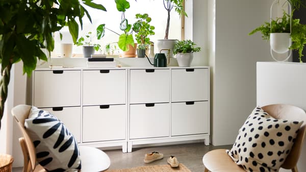 Deux range-chaussures STÄLL blancs nichés sous un appui de fenêtre garni de plantes et d’un arrosoir VATTENKRASSE.