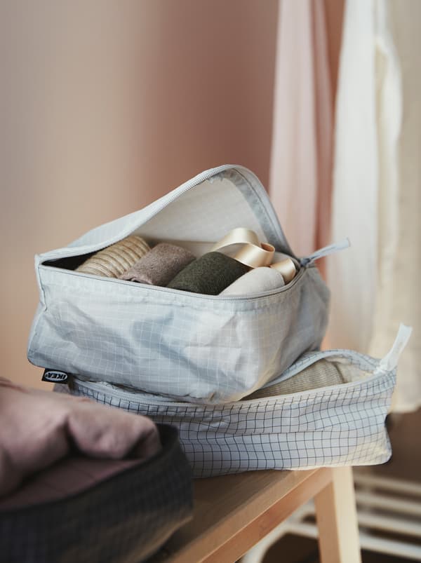 Deux sacs pour vêtements RENSARE clairs et un foncé à moitié ouverts sur un buffet, révélant des vêtements pliés avec soin.