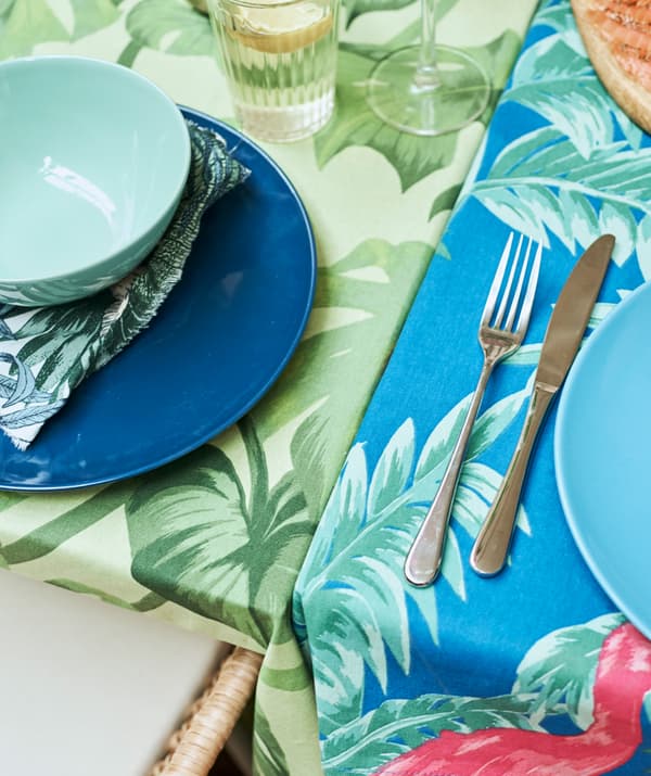 Deux tables accolées, l’une recouverte d’une nappe à imprimé de feuilles vertes, l’autre d’un tissu imprimé bleu, avec des assiettes déjà mises.