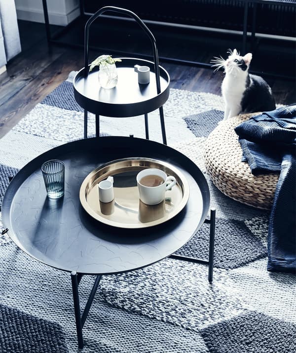 Deux tables d’appoint foncées et un repose-pieds en rotin, sur un tapis gris texturé.