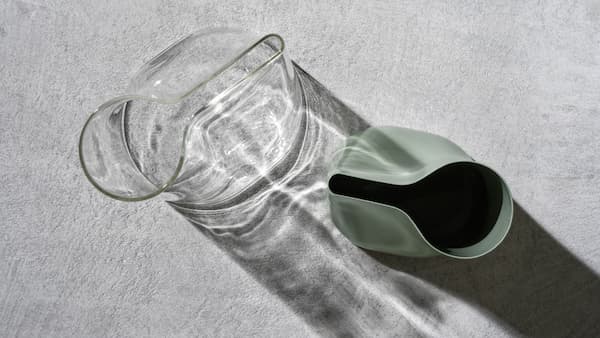 Deux vases-arrosoirs CHILIFRUKT en verre clair et en acier vert placés sur une surface en ciment gris.