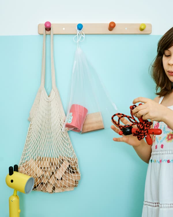 Devant un mur turquoise, une fillette tient une corde à sauter. Un sac filet contenant des blocs de bois est accroché à une patère de boutons colorés.