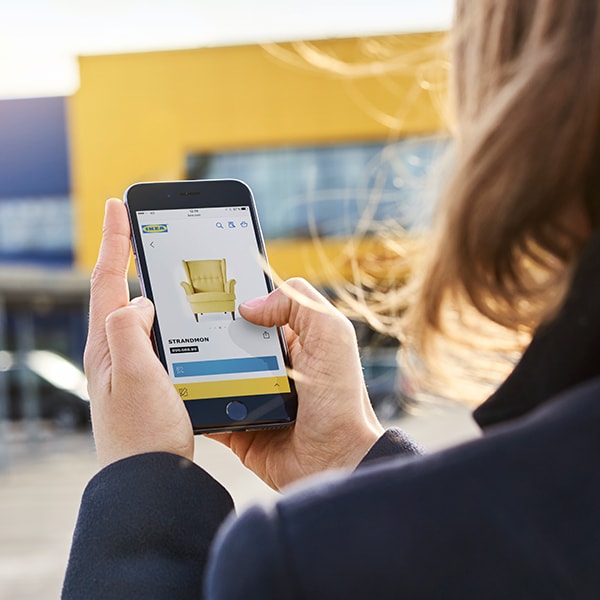 Devojka ispred robne kuće IKEA drži telefon u ruci na kojem je uključena IKEA aplikacija.