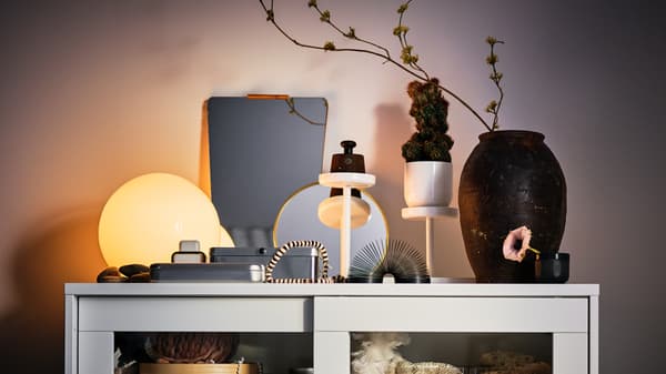 Di atas kabinet berpintu kaca SYVDE berwarna putih terdapat lampu meja FADO berwarna putih yang dinyalakan di samping berbilang item hiasan.