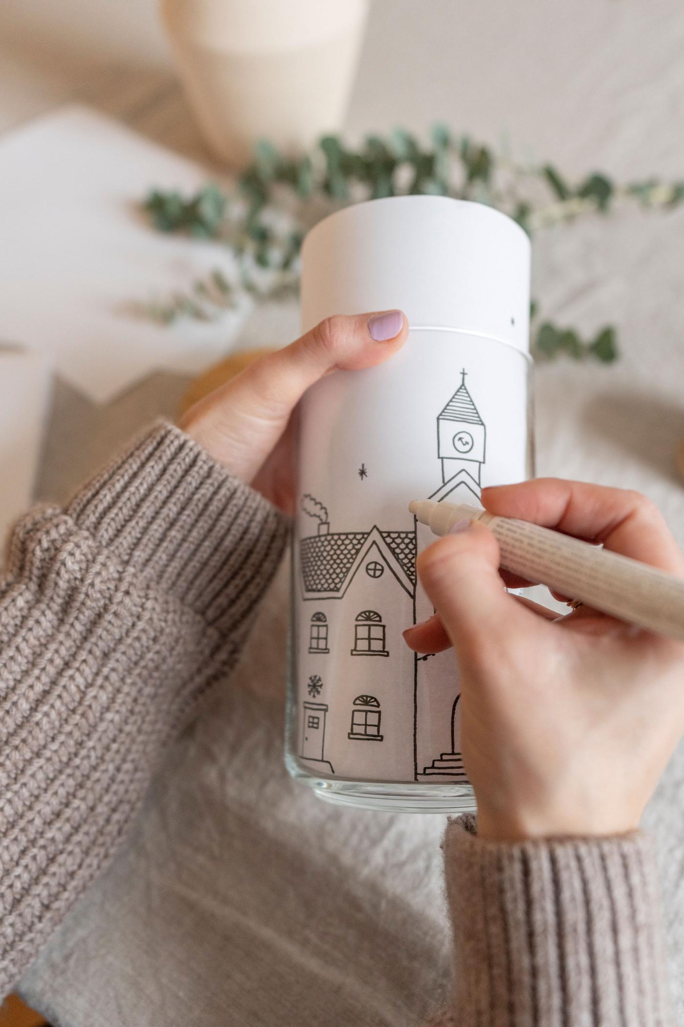 Die Malvorlage mit der winterlichen Häuserfront in einem EKLATANT Glas. Eine Hand bemalt das Glas.