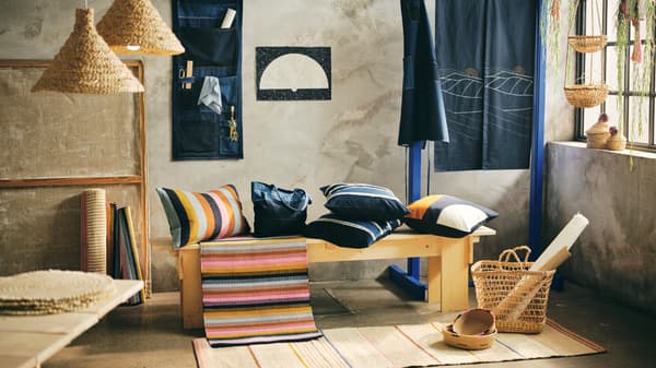 Die neue MÄVINN Kollektion von IKEA wird in einem Raum beispielhaft eingerichtet gezeigt. Ein Teppich mit bunten Streifen liegt auf einer Sitzbank und bunte Kissen liegen ebenfalls auf der Sitzbank. Die präsenten Farben auf dem Bild sind rosa, blau, gelb und unterschiedliche Beigetöne.
