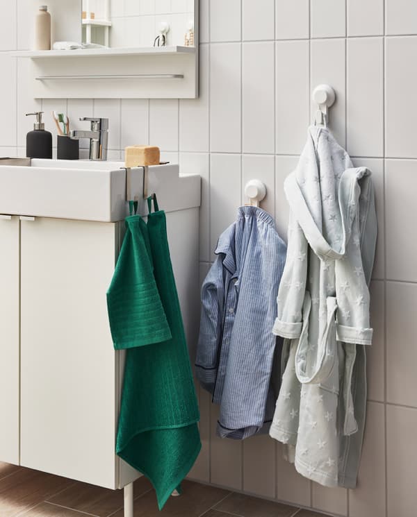 Die TISKEN Haken mit Saugnapf sind hier an der Wand neben dem Waschbecken befestigt. Daran sind ein Kinderschlafanzug und -bademantel zu sehen.