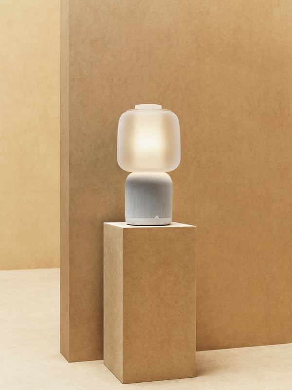 Die weiße SYMFONISK Tischleuchte steht auf einem Sockel vor einer braunen Wand.
