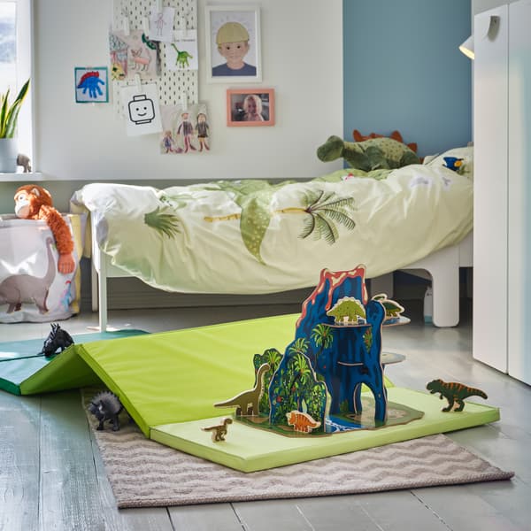 Die Welt der Dinosaurier mit Stofftierdinos ist auf einer faltbaren grünen Gymnastikmatte auf dem Fußboden nachgestellt. Im Hintergrund steht ein Bett mit grüner Bettwäsche.