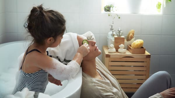 Dieťa a žena v kúpeľni. Žena sedí na podlahe a relaxuje, zatiaľ čo dieťa sa kúpe a kladie mame na oči uhorku.