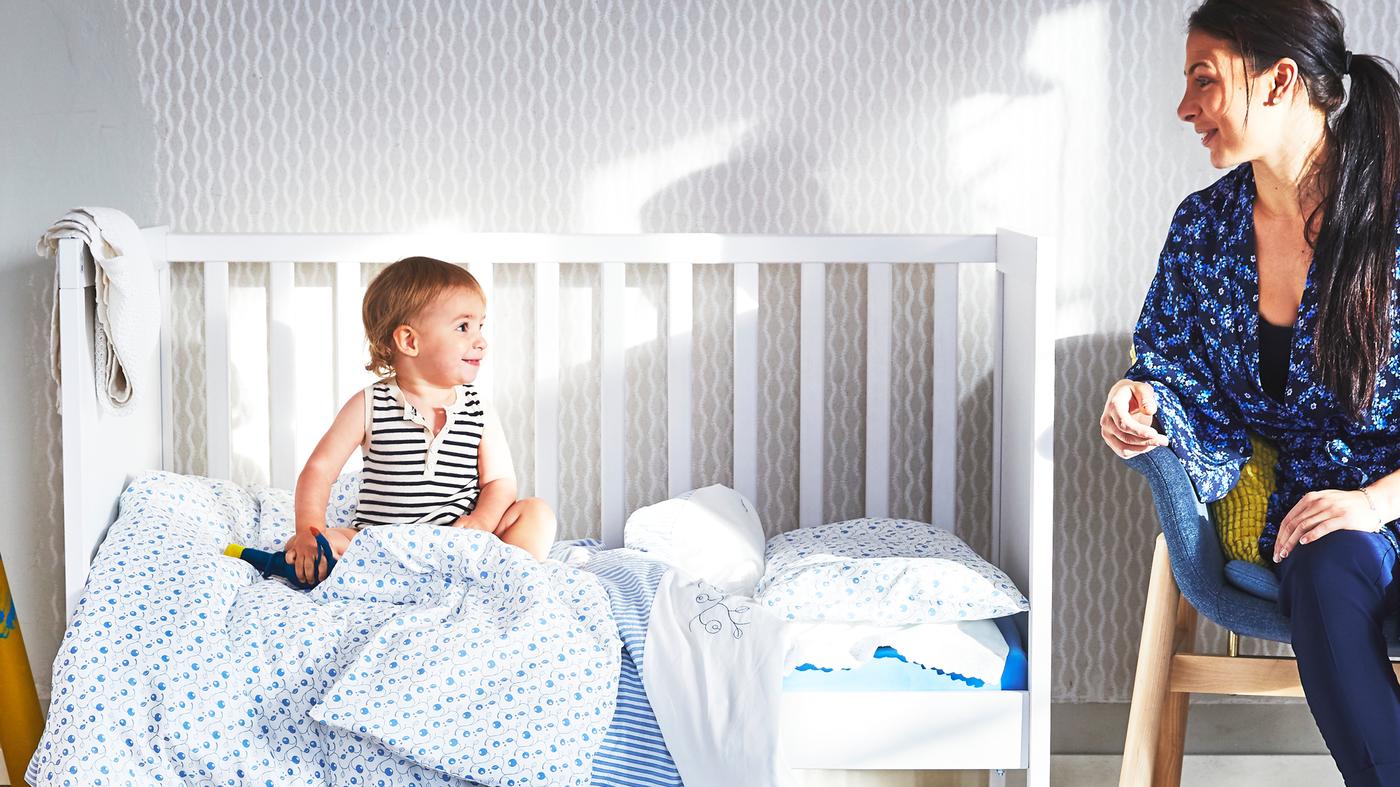 Dieťa sedí v postieľke SUNDVIK s odstránenou bočnicou a posteľnou bielizňou GULSPARV a pozerá sa na svoju mamu, ktorá sedí vedľa.