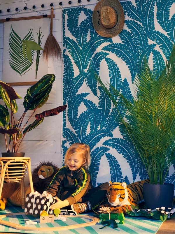 Dievča drží zabalený darček v miestnosti s rastlinami, látkou UGGLEMOTT na stene a kobercom GRACIÖS s témou džungle.