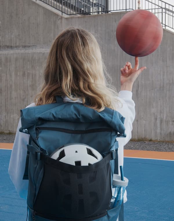 Dievča s modrým batohom VÄRLDENS s cyklistickou prilbou a fľašou vo vnútri sa hrá s basketbalovou loptou.