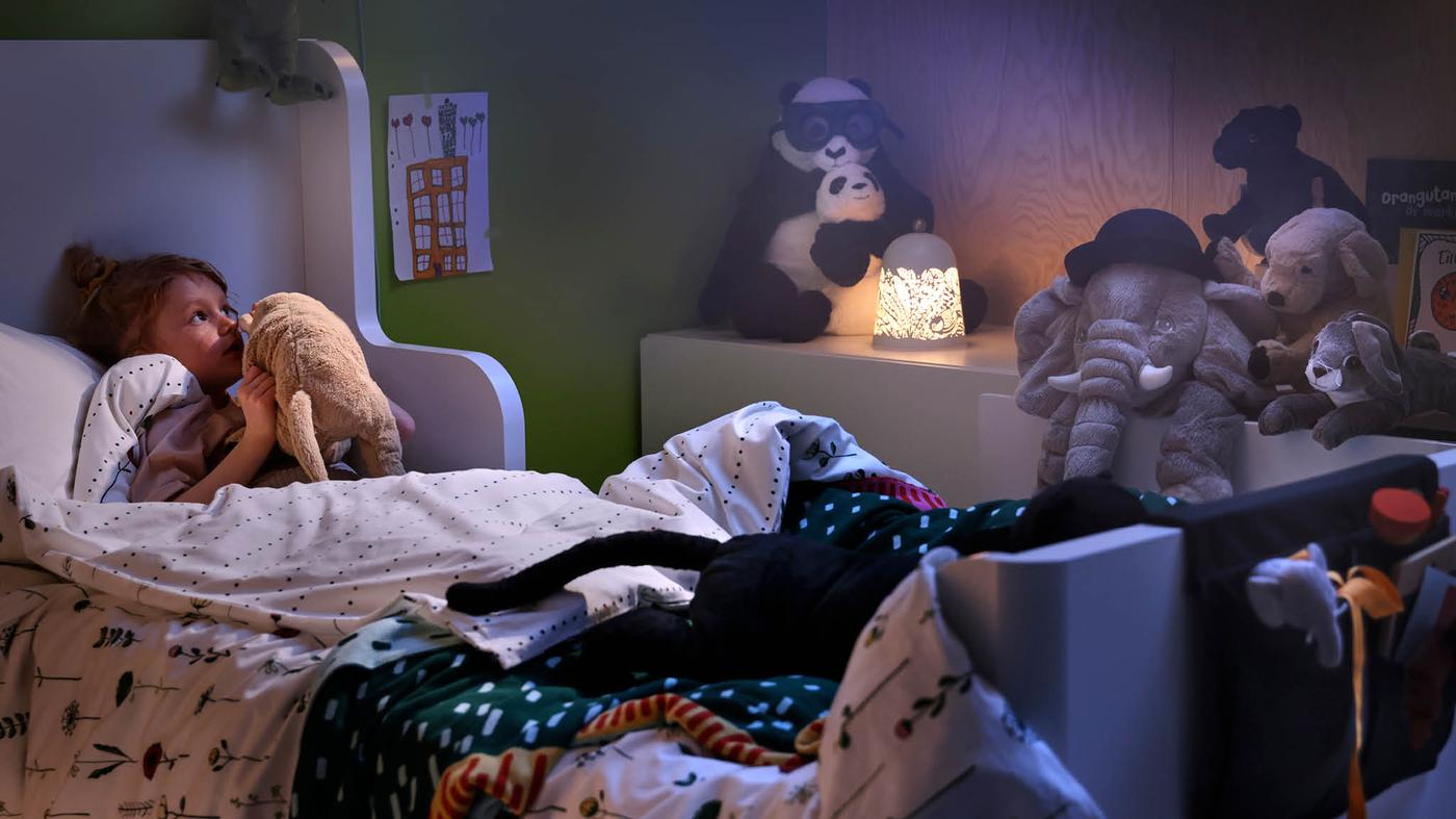 Dievčatko leží na posteli s nastaviteľnou dĺžkou BUSUNGE, v rukách drží plyšové šteniatko GOSIG GOLDEN a pozerá sa na ďalšie plyšové zvieratká na skrinke vedľa postele.