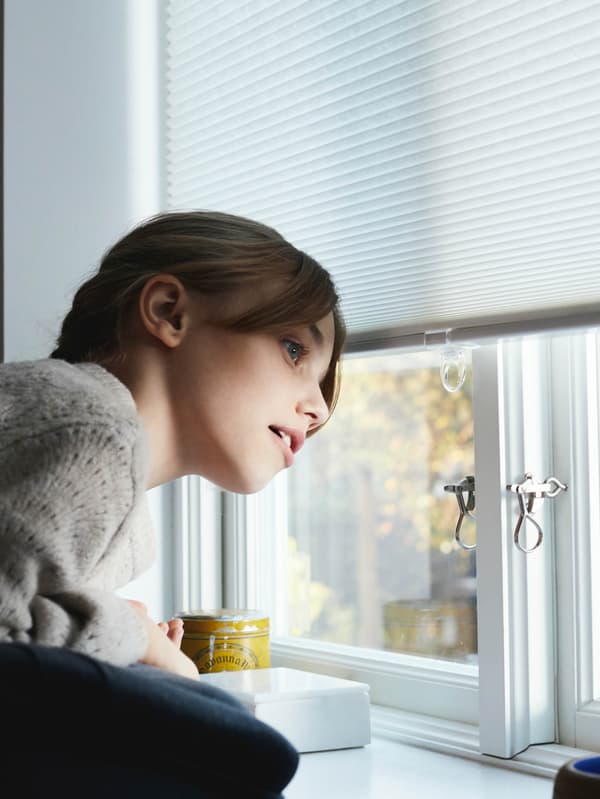 Dievčatko sa pozerá z okna, ktoré čiastočne zakrýva biela úsporná roleta HOPPVALS.