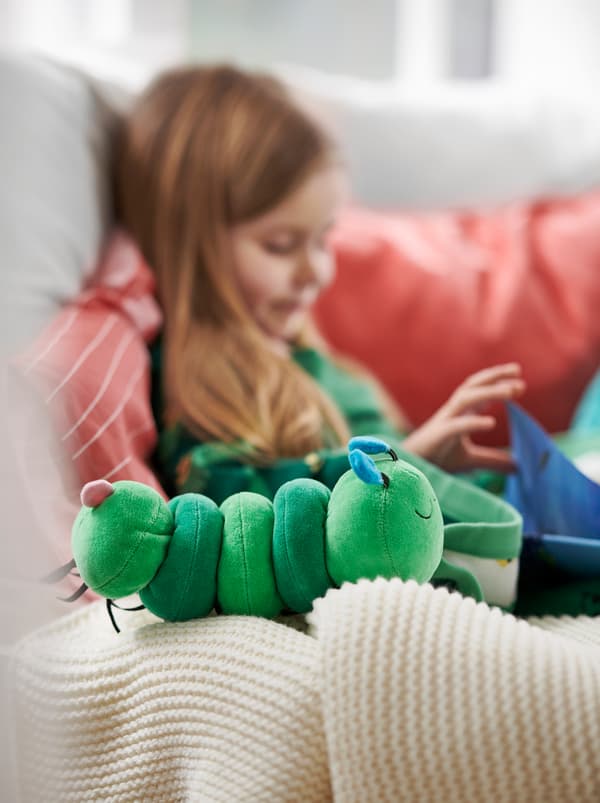 Dievčatko sedí na pohovke s červenými vankúšmi a číta knihu vedľa mäkkej zelenej hudobnej hračky KLAPPA. 