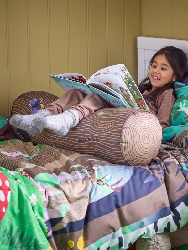 Dievčatko v posteli s obliečkami SKOGSDUVA má podopreté nohy vankúšom SKOGSDUVA a číta si knihu.