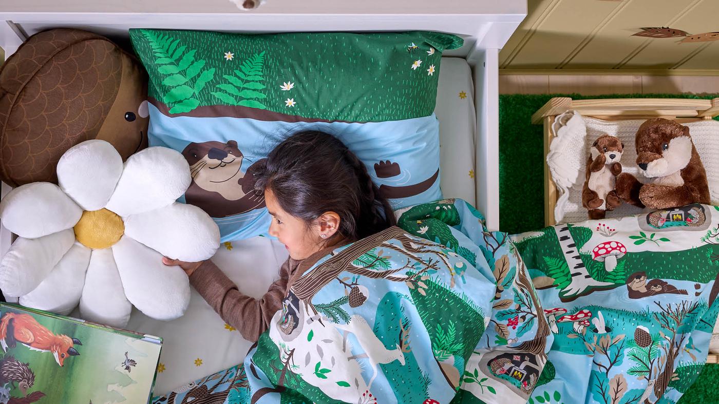 Dievčatko v posteli s obliečkami SKOGSDUVA, vedľa nej je v postieľke plyšová vydra SKOGSDUVA s mláďatkom.