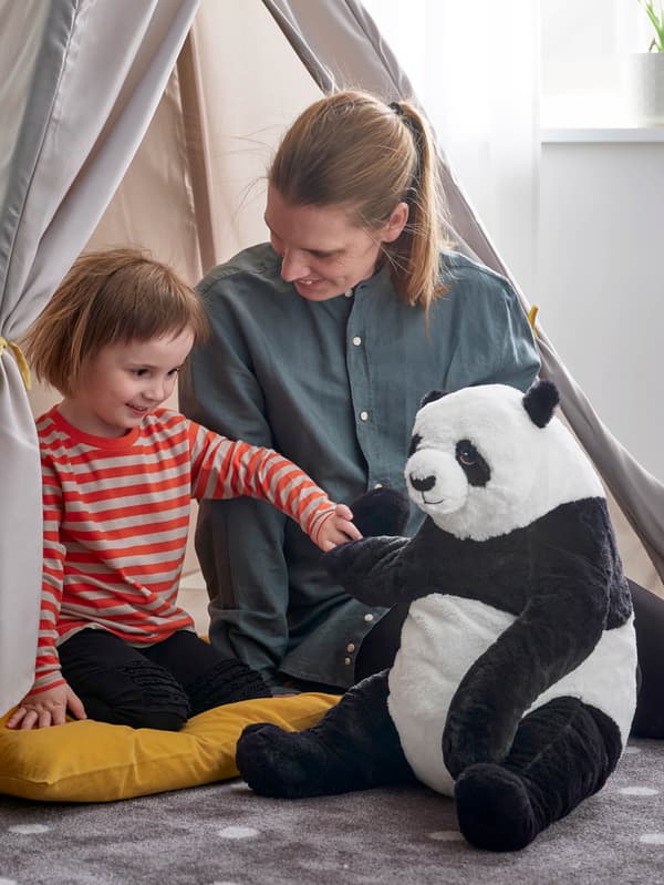 Dievčatko v pruhovanom tričku sedí na zemi na žltom vankúši vedľa mamičky a drží za labku plyšovú pandu DJUNGELSKOG.