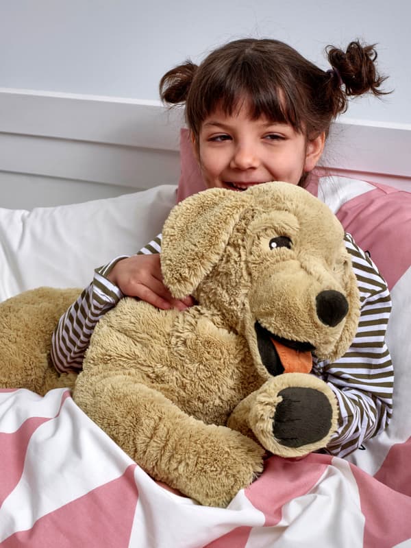 Dievčatko v pruhovanom tričku sedí v posteli a v náručí drží plyšového psa GOSIG GOLDEN.