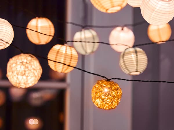 Different types of SOLVINDEN LED string lights hang on a balcony outside a window at night. The string lights are using LADDA rechargeable batteries.