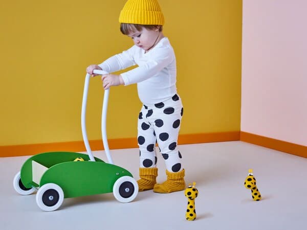 Dijete gura zelenu igračku na kotačima, nosi bijele hlače s crnim točkama i žute čarape. Oko su tri male igračke žirafe.