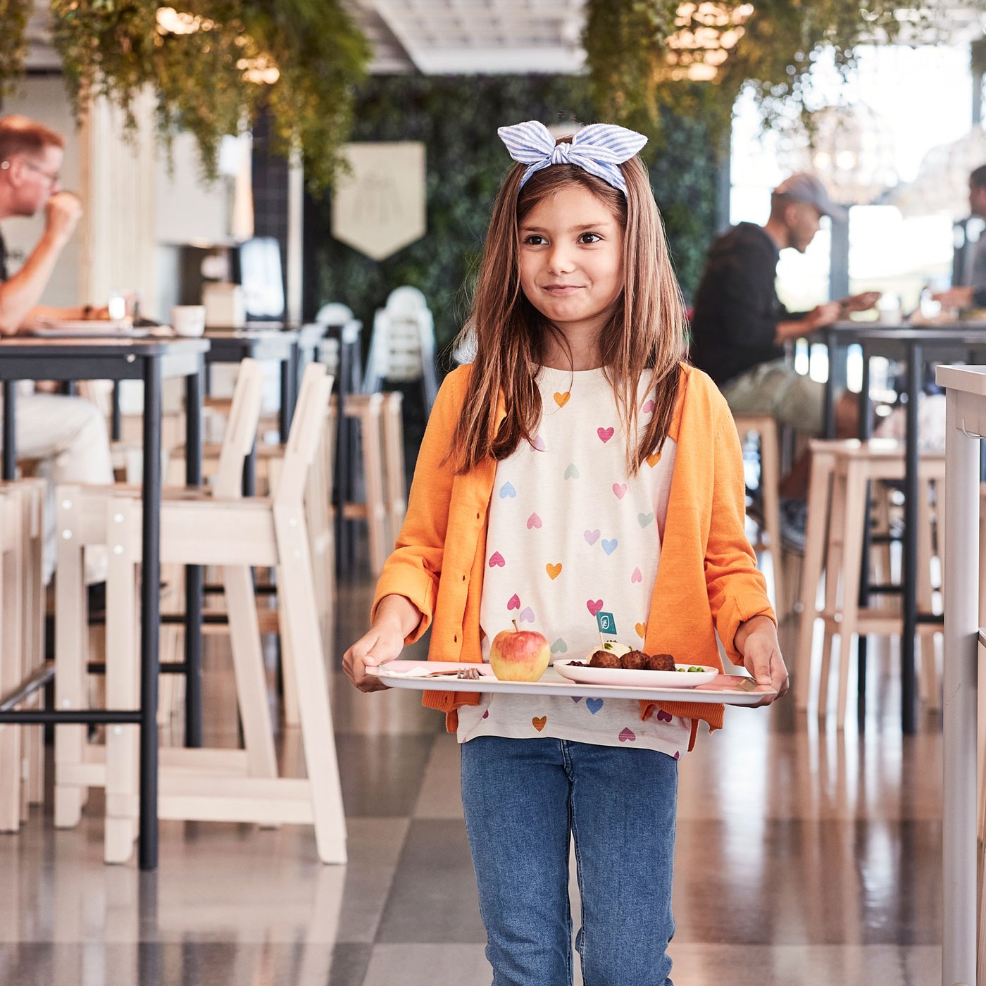 Dijete nosi pladanj s hranom u restoranu. Na pladnju su jabuka, povrće i okruglice. Dijete nosi narančasti kardigan, bijelu majicu s šarenim srcima i traperice, a na glavi ima traku s mašnom. U pozadini su stolovi i viseće biljke.