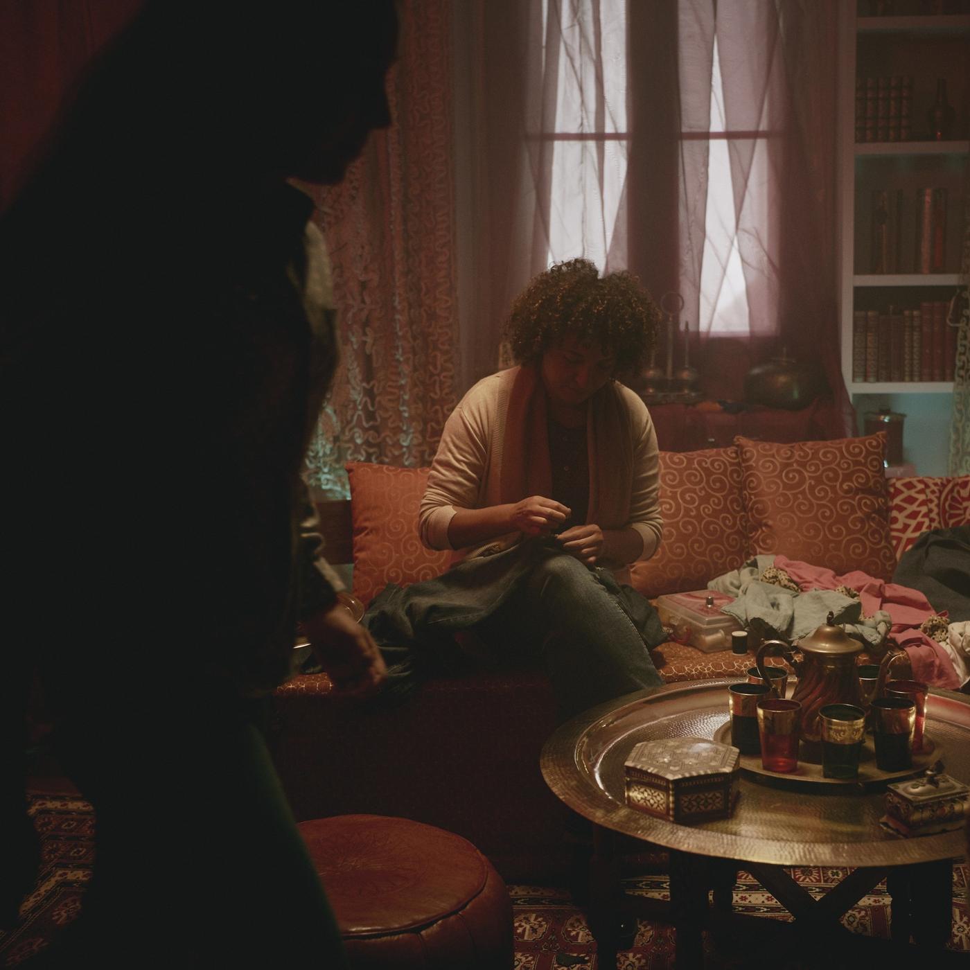 Dimly lit living room scene with a person sitting on a sofa, sewing something, while two figures stand in shadow in the foreground.