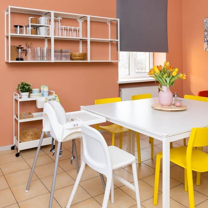 Dining area with a table surrounded by white and yellow chairs, as well as a high chair; flowers in a vase on the table and a shelf with dinnerware on the wall.