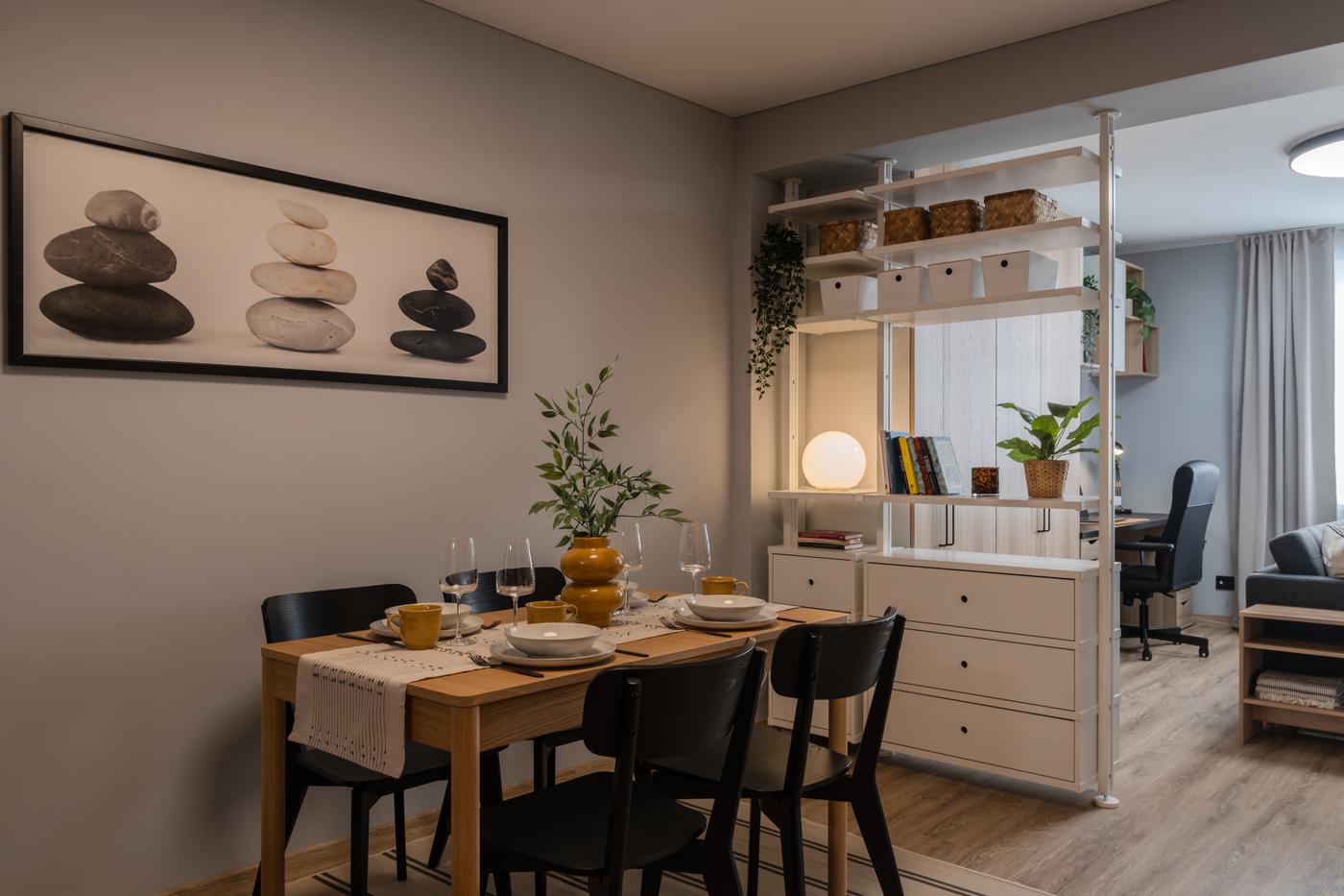 Dining area with a wooden table set for four, black chairs, and a white shelving unit with drawers separating the space from the living room.