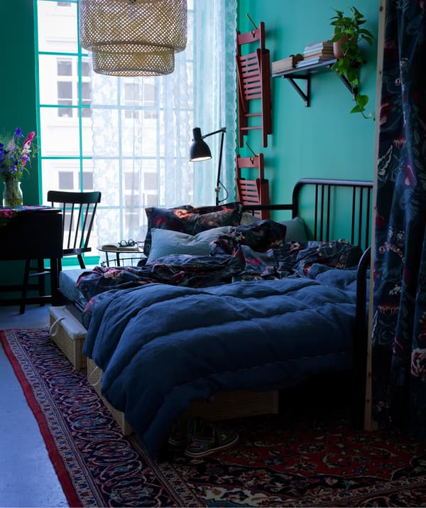 Divano letto FYRESDAL con biancheri a fiori e cuscini. Accanto, un tavolino e una sedia con scrivania. Alla parete sono appese due sedie pieghevoli rosse FRÖSVI.