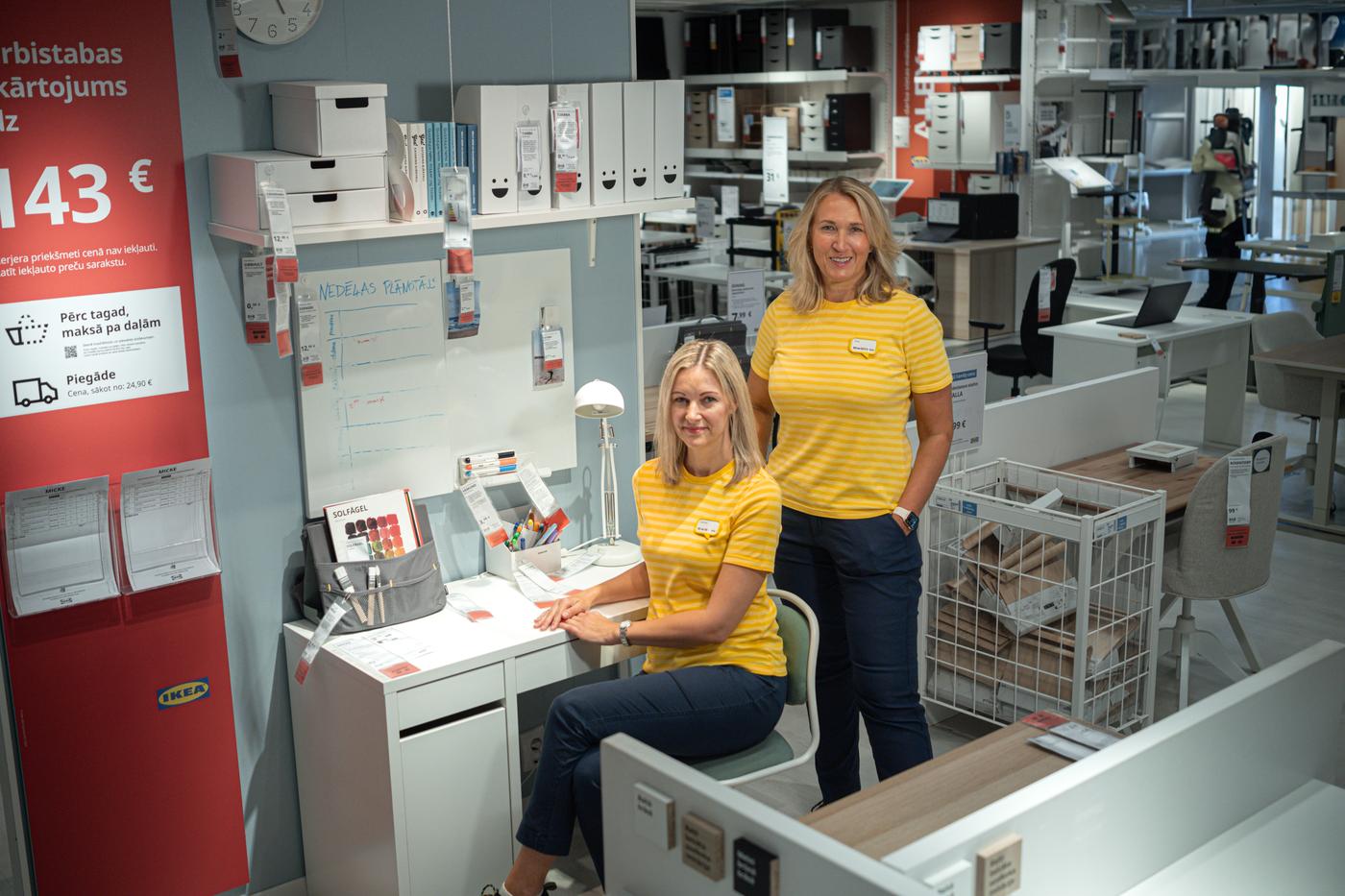 Divas IKEA darbinieces dzeltenos svītrainos kreklos pie rakstāmgalda ar plauktiem un mantu glabāšanas piederumiem.	