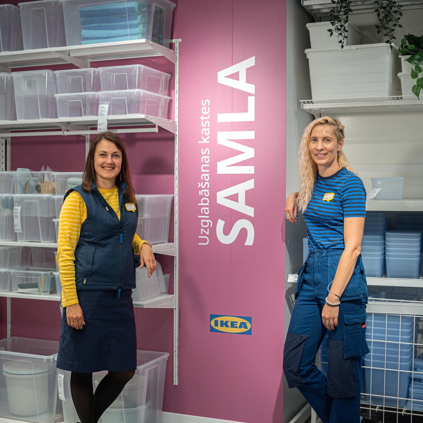 Divas IKEA darbinieces stāv pie uzglabāšanas kastēm un plauktiem; viena valkā dzeltenu svītrainu kreklu un vesti, otra – zili svītrainu kreklu.