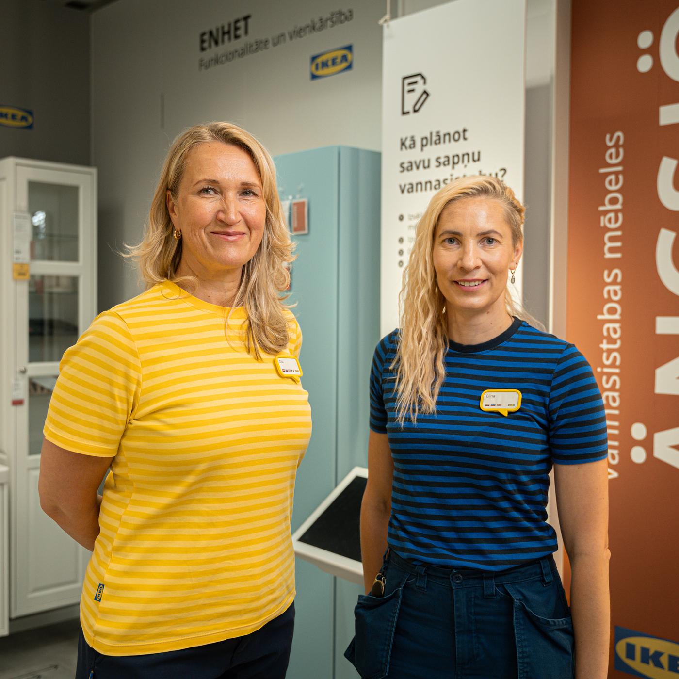 Divas IKEA formas tērpā ģērbtas ekspertes veikalā stāv un smaida kamerā, viņām aiz muguras atrodas vannasistabas mēbeļu ekspozīcija.