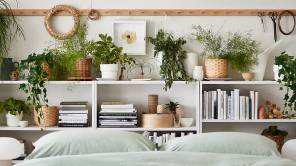 Diverse piante in vaso, fresche e artificiali, in mezzo a libri e decorazioni su librerie vicino a un letto con biancheria da letto BERGPALM - IKEA