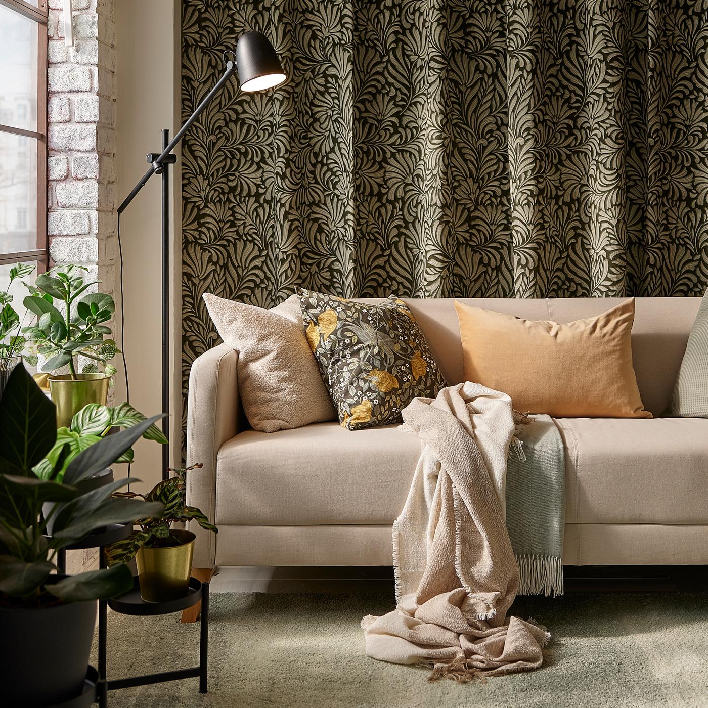 Diverse puter i en beige LINANÄS sofa på et lyst grågrønt teppe foran bladmønstrede gardiner på en vegg.