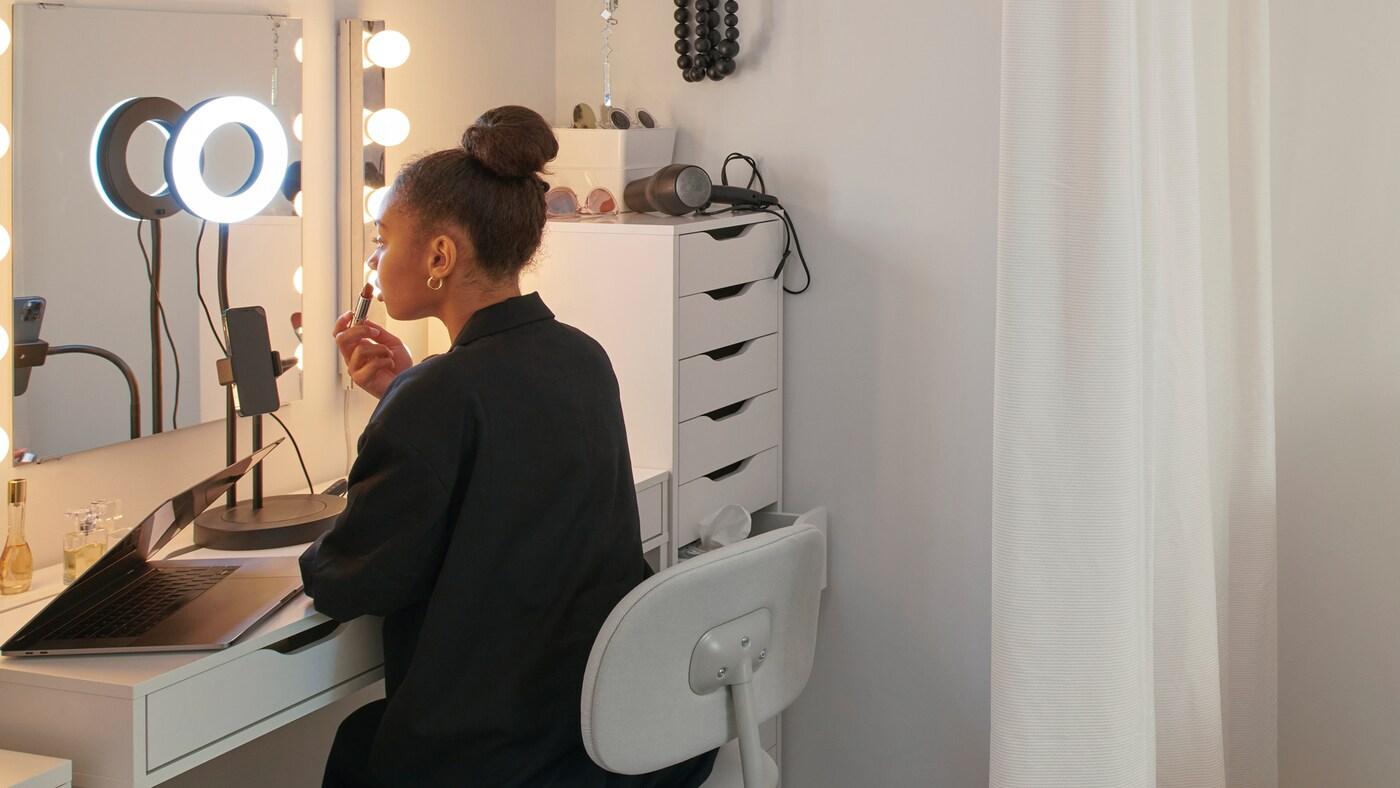 Dívka je ve svém pokoji na koleji na otočné židli BLECKBERGET u stolu MICKE. Nanáší si make-up a svítí si kruhovým světlem LÅNESPELARE.