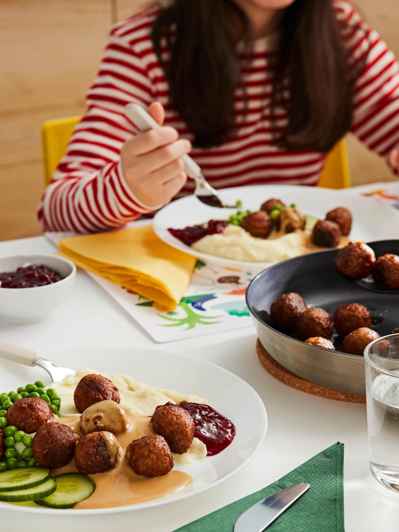 Dívka v červeno-bílo pruhovaném tričku sedí u stolu nad talířem masových kuliček HUVUDROLL  s bramborovou kaší a brusinkovým džemem, před ní je pánev s kuličkami a talíř se další porcí kuliček, kaše, omáčky, džemu a hrášku.